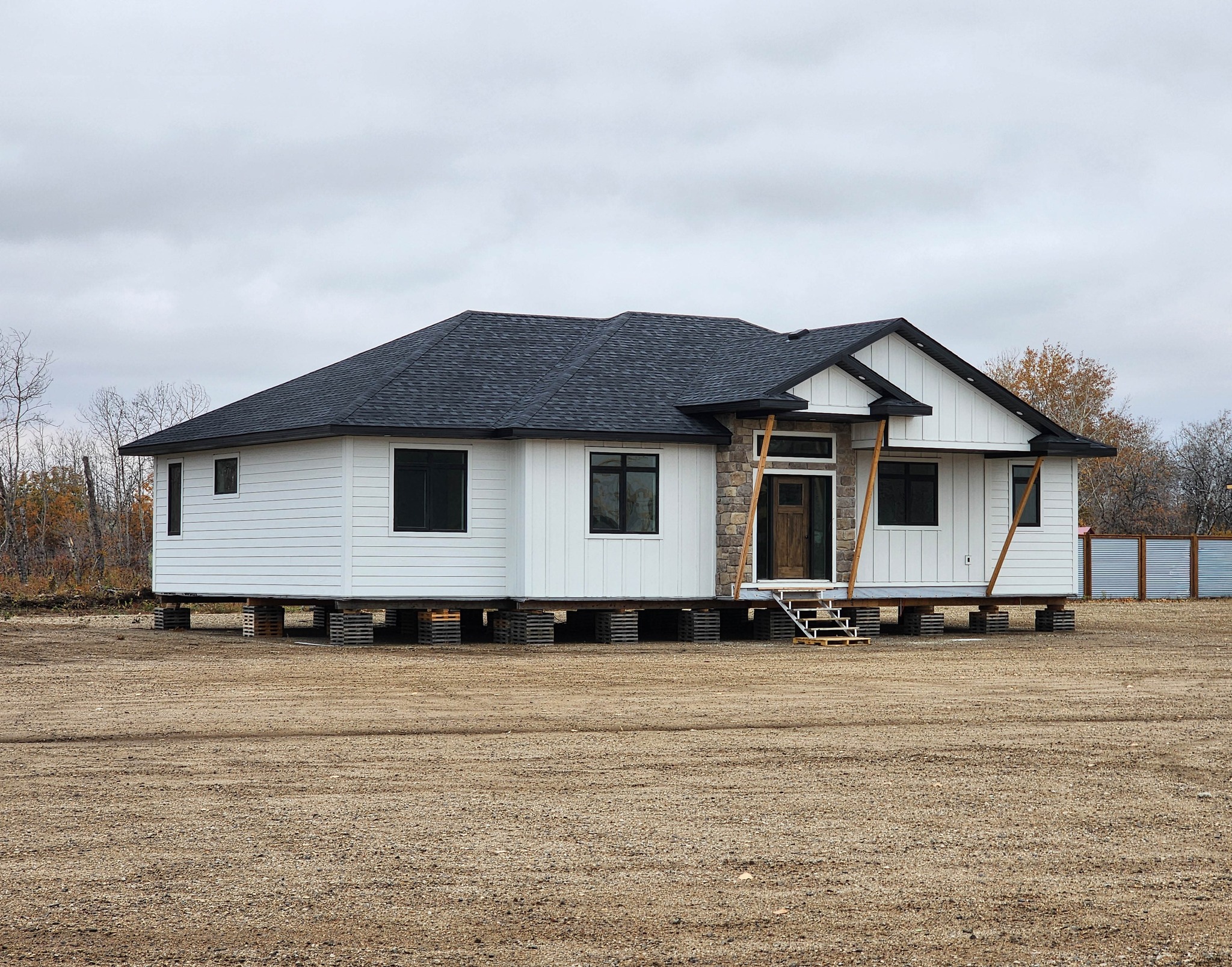 Single-story white house on blocks in a barren yard. Black roof, stone accent, and wooden scaffolding. Overcast sky, sparse trees, and a quiet, unfinished mood.