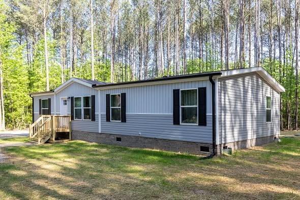 Single-story, gray-paneled mobile home with black shutters, nestled among tall pine trees. Sunlight filters through, creating a serene atmosphere.