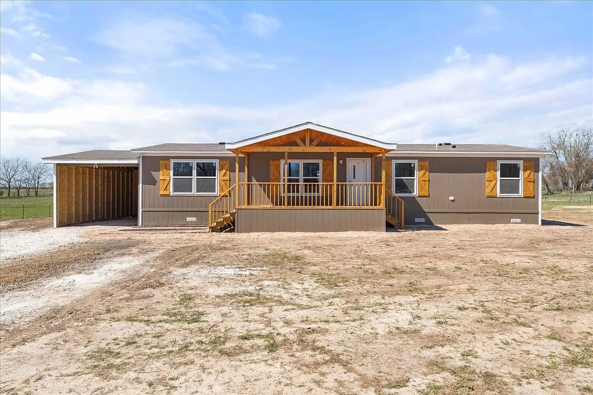 Single-story beige house with wooden shutters and a front porch. It has a carport on the left. The setting is open land with a bright sky.