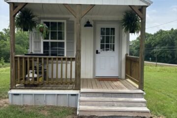 A small, white, rustic cabin with a wooden porch and steps, adorned with green hanging plants, set in a lush grassy field under a cloudy sky.