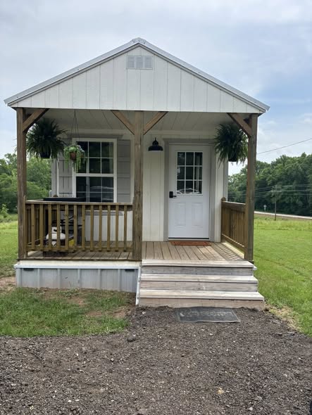 A small, white, rustic cabin with a wooden porch and steps, adorned with green hanging plants, set in a lush grassy field under a cloudy sky.