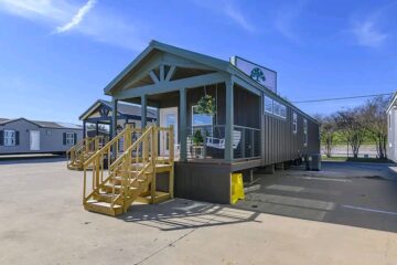 A tiny house with a porch is displayed under a clear blue sky. The porch has wooden steps and a hanging flower basket, conveying a welcoming feel.