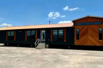 A modern, modular home is set on a gravel lot under a clear blue sky. It features a dark exterior with wood accents and large windows, evoking a sleek, contemporary feel.