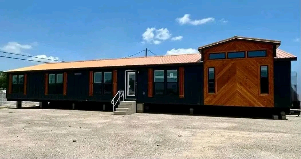 A modern, modular home is set on a gravel lot under a clear blue sky. It features a dark exterior with wood accents and large windows, evoking a sleek, contemporary feel.