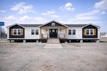 A modern, single-story manufactured home. The exterior features dark siding with white panels and wooden shutters, flanked by double stairs leading to the black front door. The sky is clear with scattered clouds.