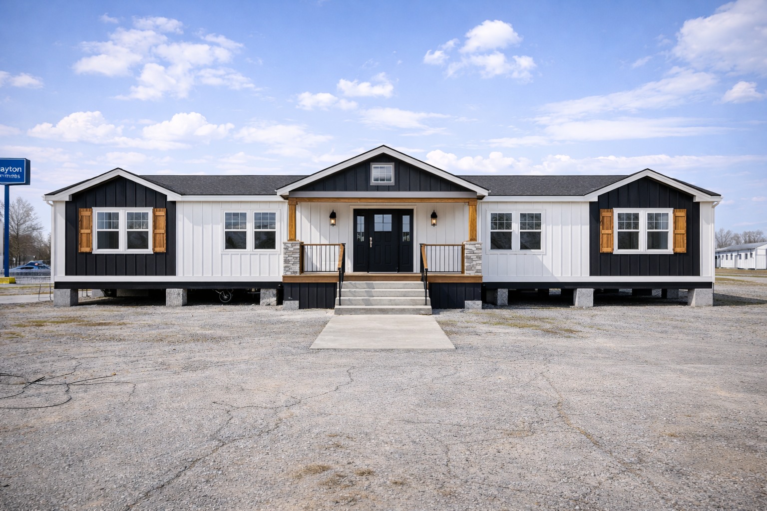 A modern, single-story manufactured home. The exterior features dark siding with white panels and wooden shutters, flanked by double stairs leading to the black front door. The sky is clear with scattered clouds.