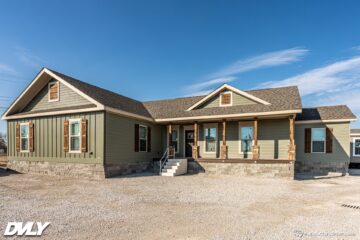 Single-story modular home with green siding and stone base, featuring a covered porch with wooden pillars. The sky is clear and the setting is calm.