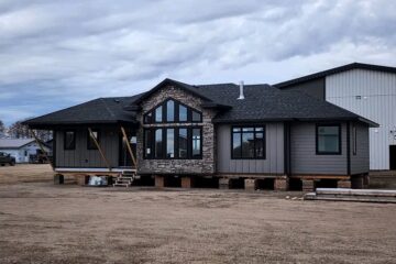 Single-story house raised on blocks, with gray siding and stone accents. Large windows, wooden supports, under a cloudy sky. Rural setting.