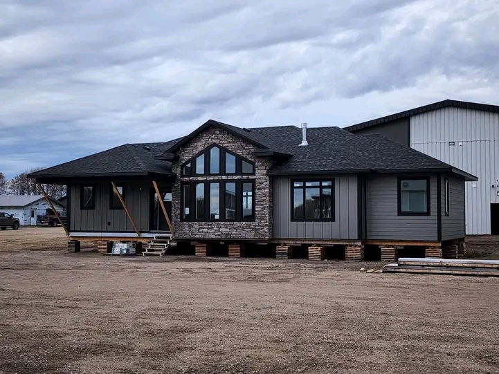 Single-story house raised on blocks, with gray siding and stone accents. Large windows, wooden supports, under a cloudy sky. Rural setting.