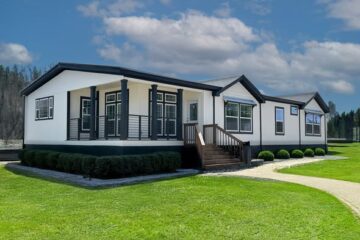 A modern white modular home with black trim sits on a well-manicured lawn under a blue sky with clouds. Its large windows and steps convey a welcoming feel.