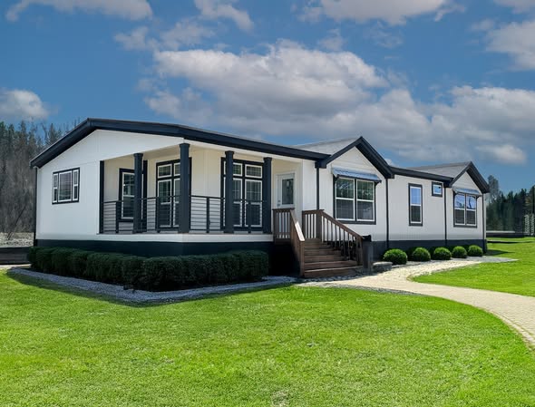 A modern white modular home with black trim sits on a well-manicured lawn under a blue sky with clouds. Its large windows and steps convey a welcoming feel.