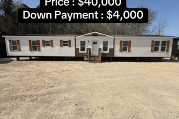 Long mobile home with beige siding and dark wooden shutters in a gravel lot. Text overlay shows price: $40,000 and down payment: $4,000. Bright, sunny day.