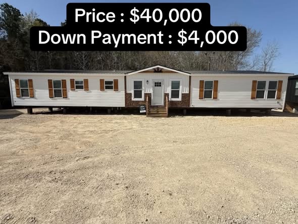 Long mobile home with beige siding and dark wooden shutters in a gravel lot. Text overlay shows price: $40,000 and down payment: $4,000. Bright, sunny day.