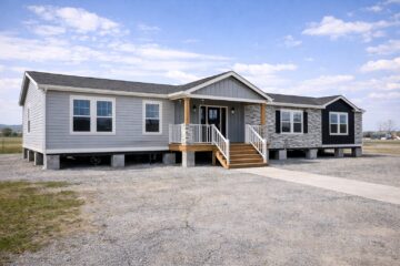 Single-story modular home with gray siding, stone accents, and a wooden porch. Set on a gravel lot under a clear, blue sky, conveying simplicity and tranquility.