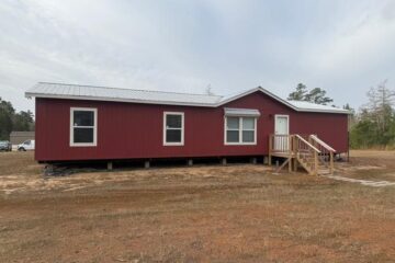 A red, single-story modular home with a white door and windows, set on a grassy field under an overcast sky. Wooden steps lead up to the entrance.