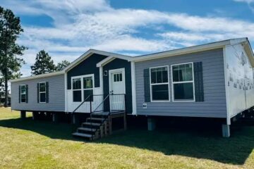A modern, single-story prefabricated home with gray siding and white trim stands on green grass under a bright, blue sky with wispy clouds.