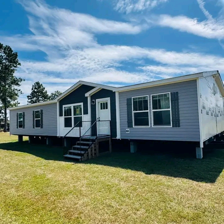 A modern, single-story prefabricated home with gray siding and white trim stands on green grass under a bright, blue sky with wispy clouds.