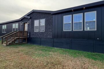 Single-story, dark gray manufactured home with three-paneled windows and stone accent, nestled on a grassy lawn under an overcast sky.