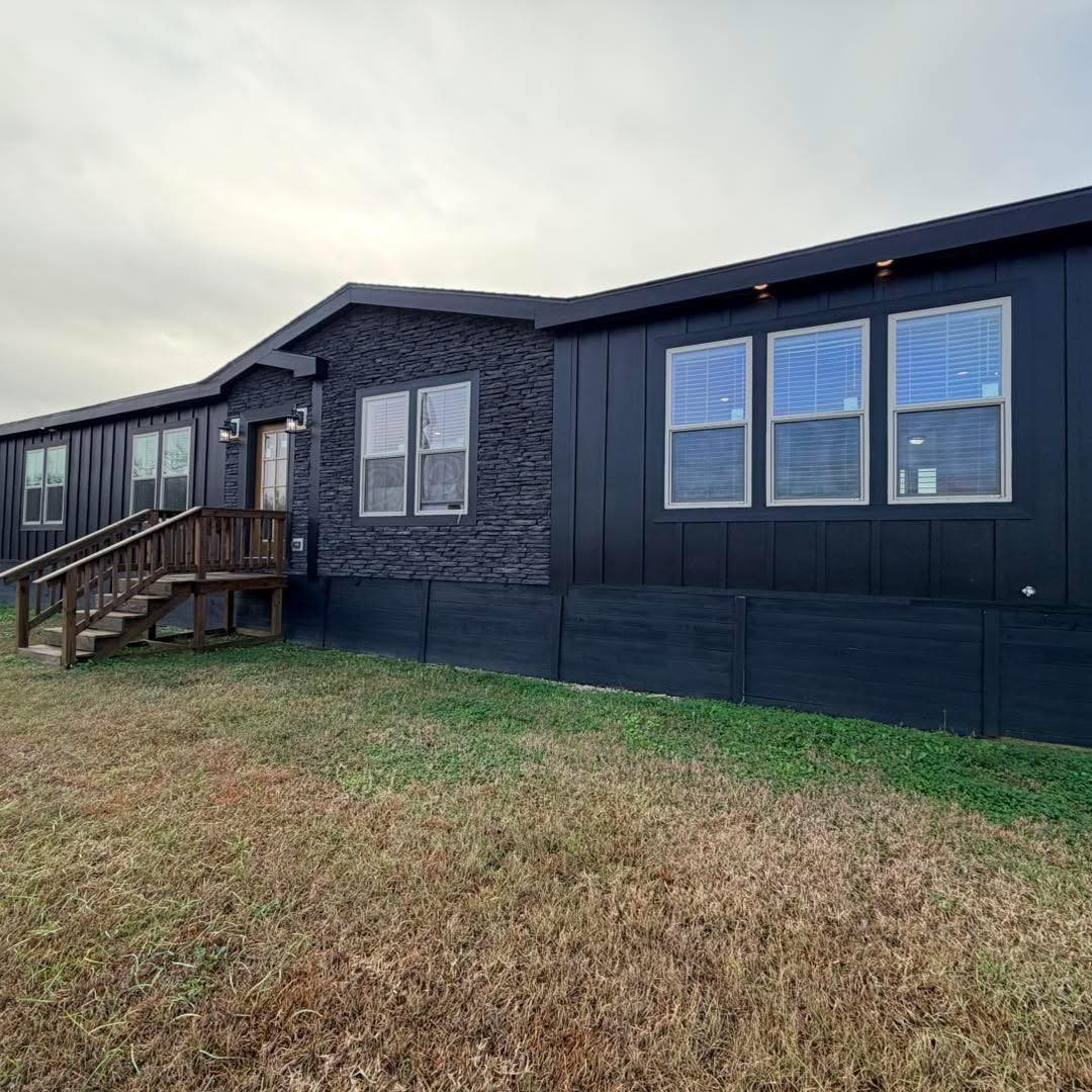 Single-story, dark gray manufactured home with three-paneled windows and stone accent, nestled on a grassy lawn under an overcast sky.