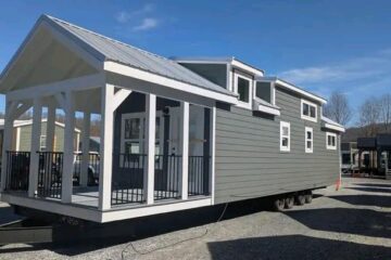A modern gray tiny house on wheels with a white-trimmed covered porch and multiple windows, set in a sunny landscape with trees in the background.