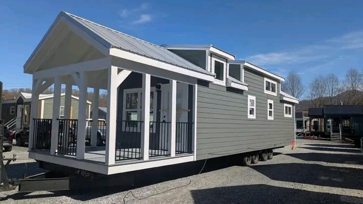 A modern gray tiny house on wheels with a white-trimmed covered porch and multiple windows, set in a sunny landscape with trees in the background.