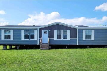 A blue and white mobile home sits on a grassy field under a clear blue sky. It features large windows and a small porch with steps leading up.