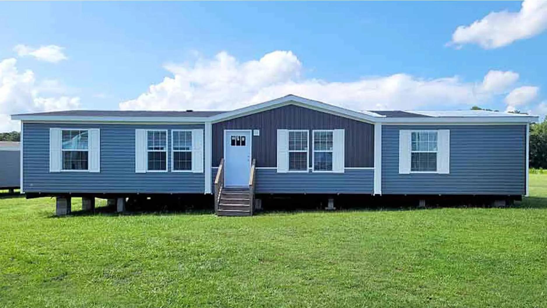 A blue and white mobile home sits on a grassy field under a clear blue sky. It features large windows and a small porch with steps leading up.