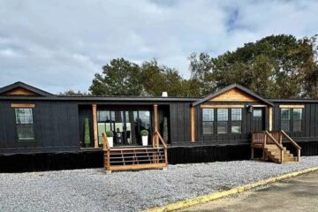 A modern black mobile home with wooden accents, featuring two small porches, stands on a gravel area. Trees are in the background under a cloudy sky.