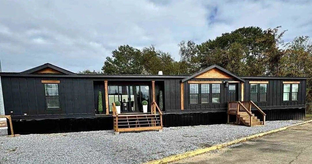 A modern black mobile home with wooden accents, featuring two small porches, stands on a gravel area. Trees are in the background under a cloudy sky.