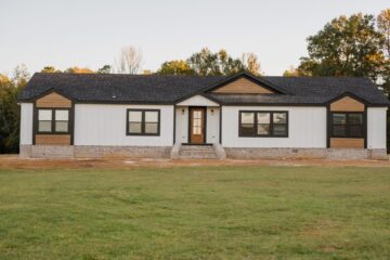 A modern single-story house with white siding, black trim, and wooden accents. Set against a backdrop of trees with a clear sky and a grassy lawn in the foreground.