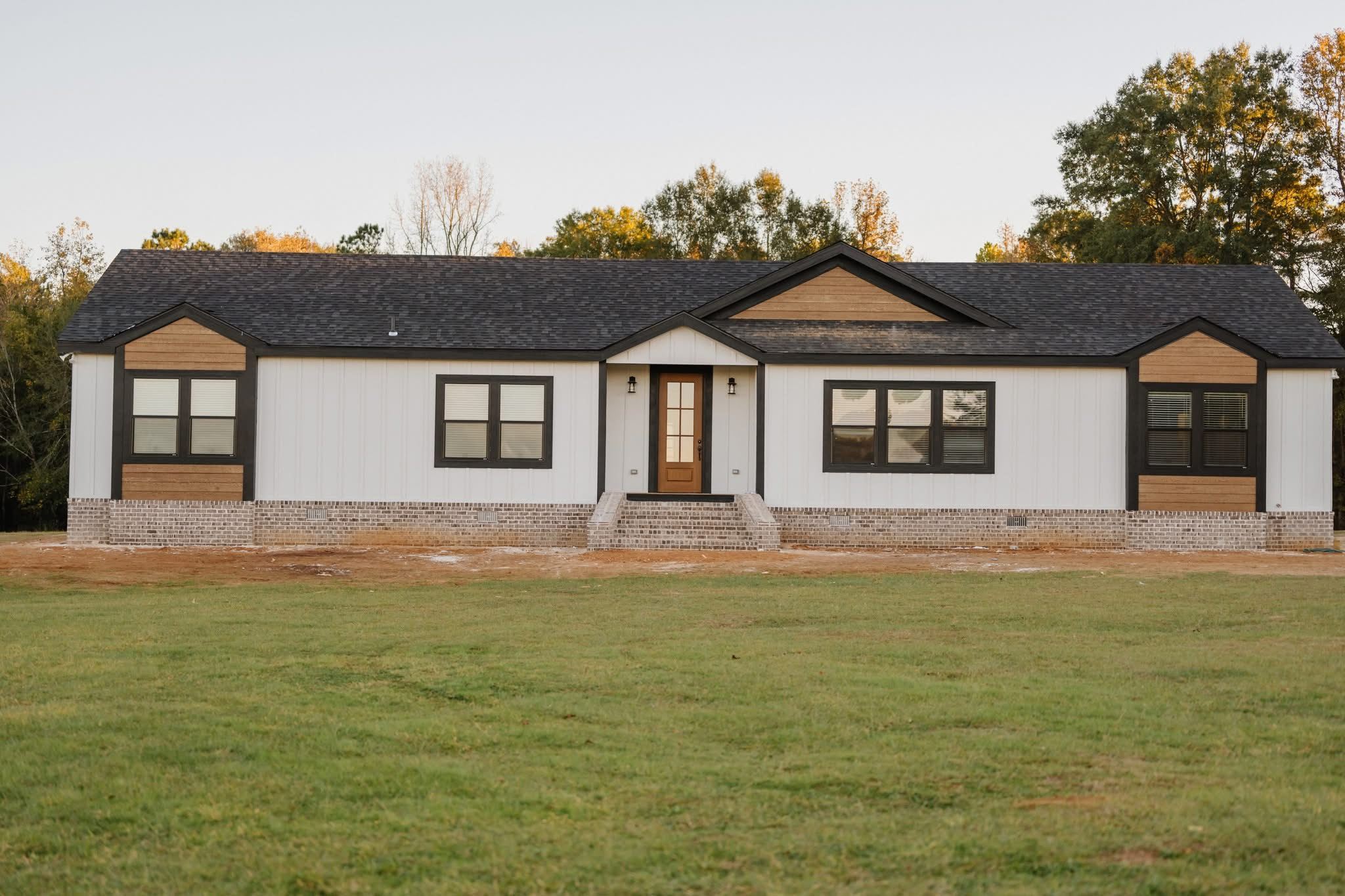 A modern single-story house with white siding, black trim, and wooden accents. Set against a backdrop of trees with a clear sky and a grassy lawn in the foreground.