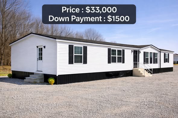 A white manufactured home with black shutters sits on a gravel lot. Text overlays indicate a price of $33,000 and a down payment of $1,500. Trees are in the background.
