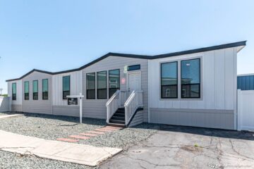 Modern white manufactured home with black trim, featuring large windows and a central front door accessed by stairs. Neat gravel yard and clear blue sky.