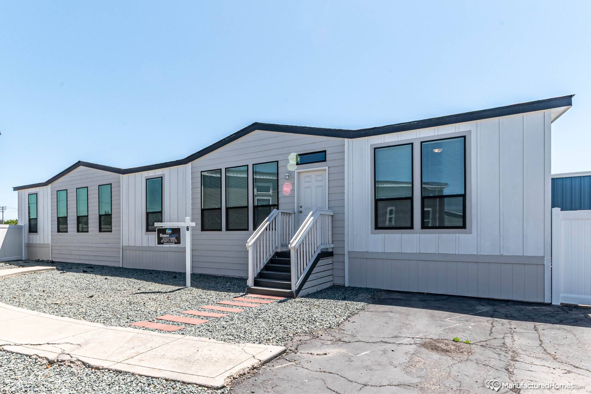 Modern white manufactured home with black trim, featuring large windows and a central front door accessed by stairs. Neat gravel yard and clear blue sky.