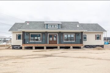 Single-story house on a dirt lot, featuring light siding, a dark central section with a wooden porch and double doors, under an overcast sky.