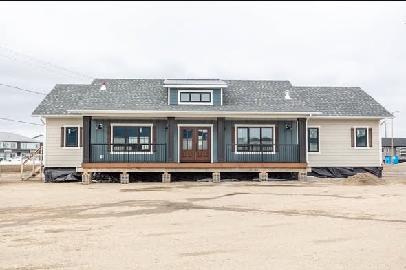 Single-story house on a dirt lot, featuring light siding, a dark central section with a wooden porch and double doors, under an overcast sky.