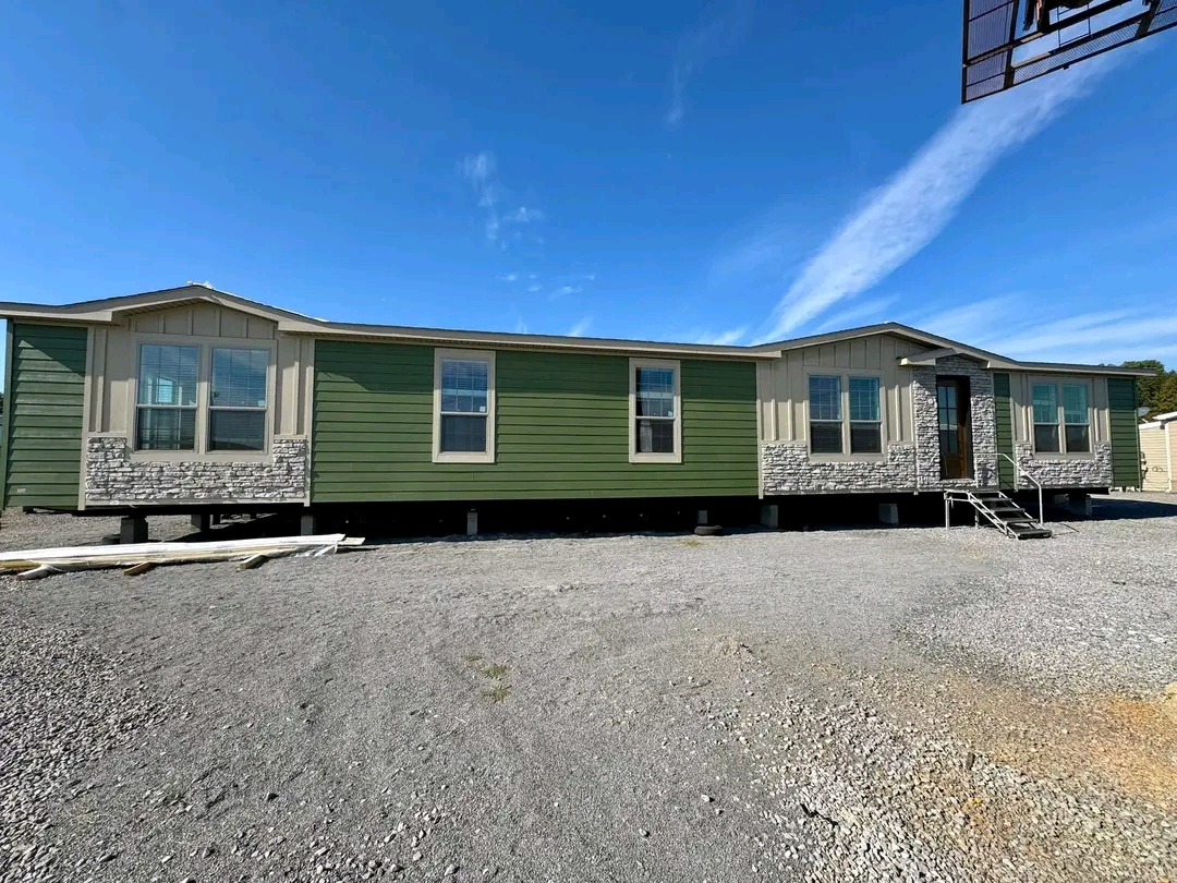 A green manufactured home with beige stone accents sits on a gravel lot under a clear blue sky. Large windows and a small metal staircase are visible.