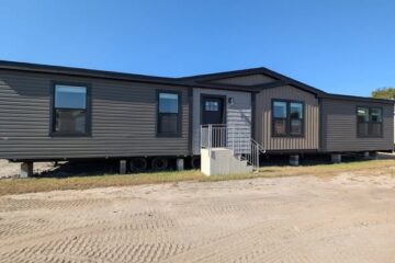 A brown manufactured home with three sections, mounted on wheels, sits on a dirt lot under a clear blue sky, giving a sense of simplicity and functionality.