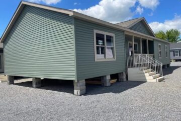 A green mobile home elevated on concrete blocks with white trim, front porch, and steps. The sky is clear with scattered clouds, creating a calm atmosphere.