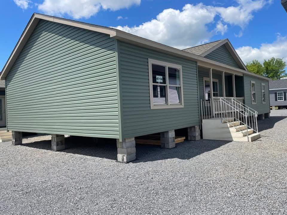 A green mobile home elevated on concrete blocks with white trim, front porch, and steps. The sky is clear with scattered clouds, creating a calm atmosphere.