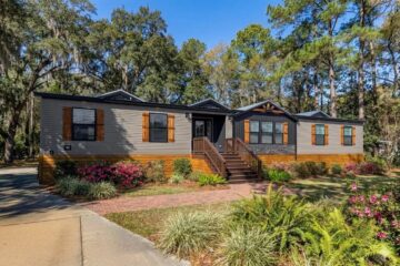 Single-story modular home with gray siding and wood shutters, surrounded by lush green trees and vibrant flowering plants, evoking a welcoming atmosphere.