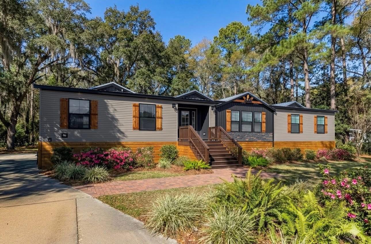 Single-story modular home with gray siding and wood shutters, surrounded by lush green trees and vibrant flowering plants, evoking a welcoming atmosphere.