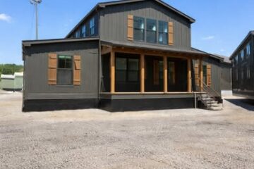 A modern black cabin with wooden shutters and a porch stands under a clear blue sky. The gravel ground and empty surroundings add a rustic vibe.