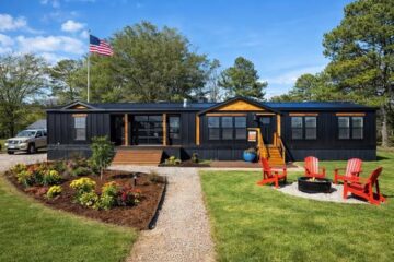 Black modular home with wood accents, surrounded by green lawn and colorful flowers. Red chairs encircle a fire pit. An American flag waves nearby.