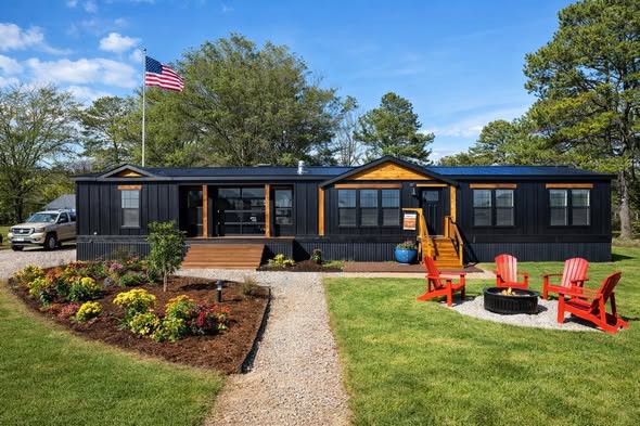 Black modular home with wood accents, surrounded by green lawn and colorful flowers. Red chairs encircle a fire pit. An American flag waves nearby.