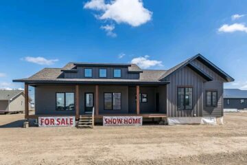 Single-story show home with gray siding, covered porch, and a "For Sale" sign. Bright blue sky with scattered clouds creates an inviting atmosphere.