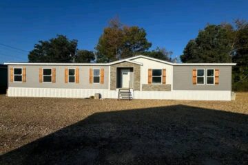 A single-story mobile home with light gray siding and brown shutters sits under a clear blue sky, surrounded by trees. The mood is serene and inviting.