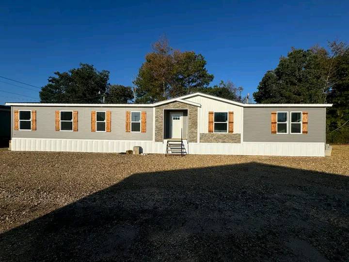 A single-story mobile home with light gray siding and brown shutters sits under a clear blue sky, surrounded by trees. The mood is serene and inviting.