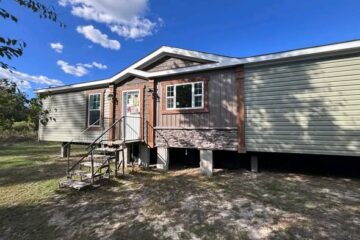Single-story house with light green siding and stone accents. A small porch with steps leads to the front door. Clear blue sky with some clouds.