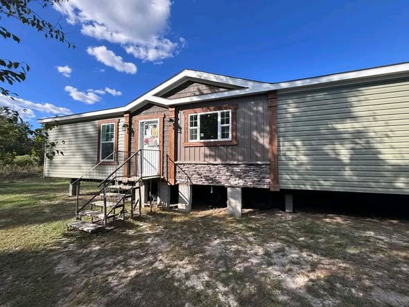 Single-story house with light green siding and stone accents. A small porch with steps leads to the front door. Clear blue sky with some clouds.